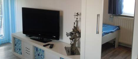 A room with a television on a white cabinet, a bed with a blue mattress, and a window with curtains.