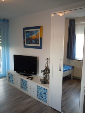 Une pièce avec un téléviseur posé sur un meuble blanc, un lit avec un matelas bleu et une fenêtre avec des rideaux.
