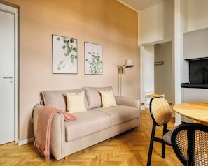 Una sala moderna con un sofá, dos láminas botánicas enmarcadas, una lámpara de pie y una zona de comedor con una mesa y sillas.