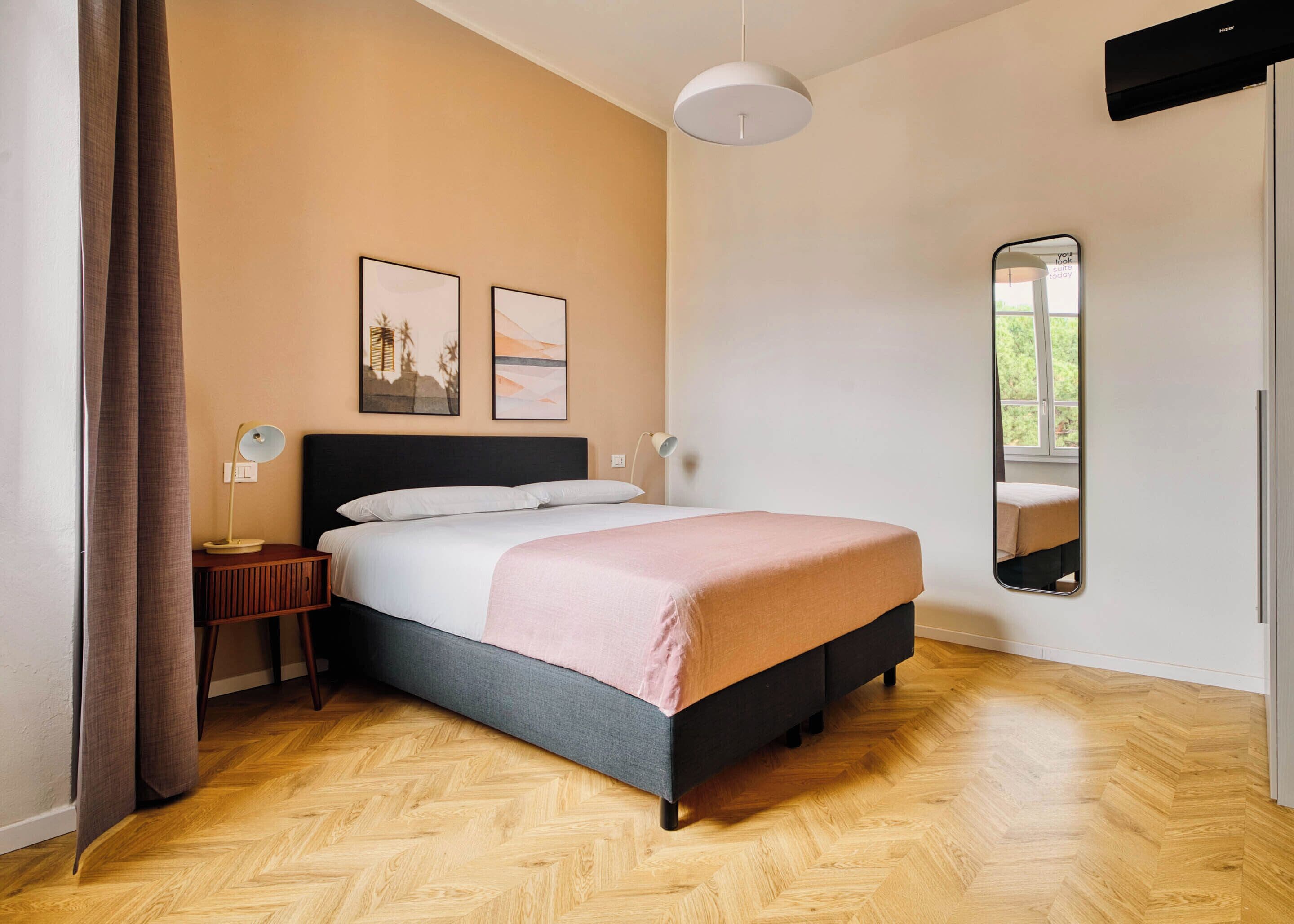 Un dormitorio con cama, mesita de noche, lámpara, espejo y dos cuadros colgados en la pared.