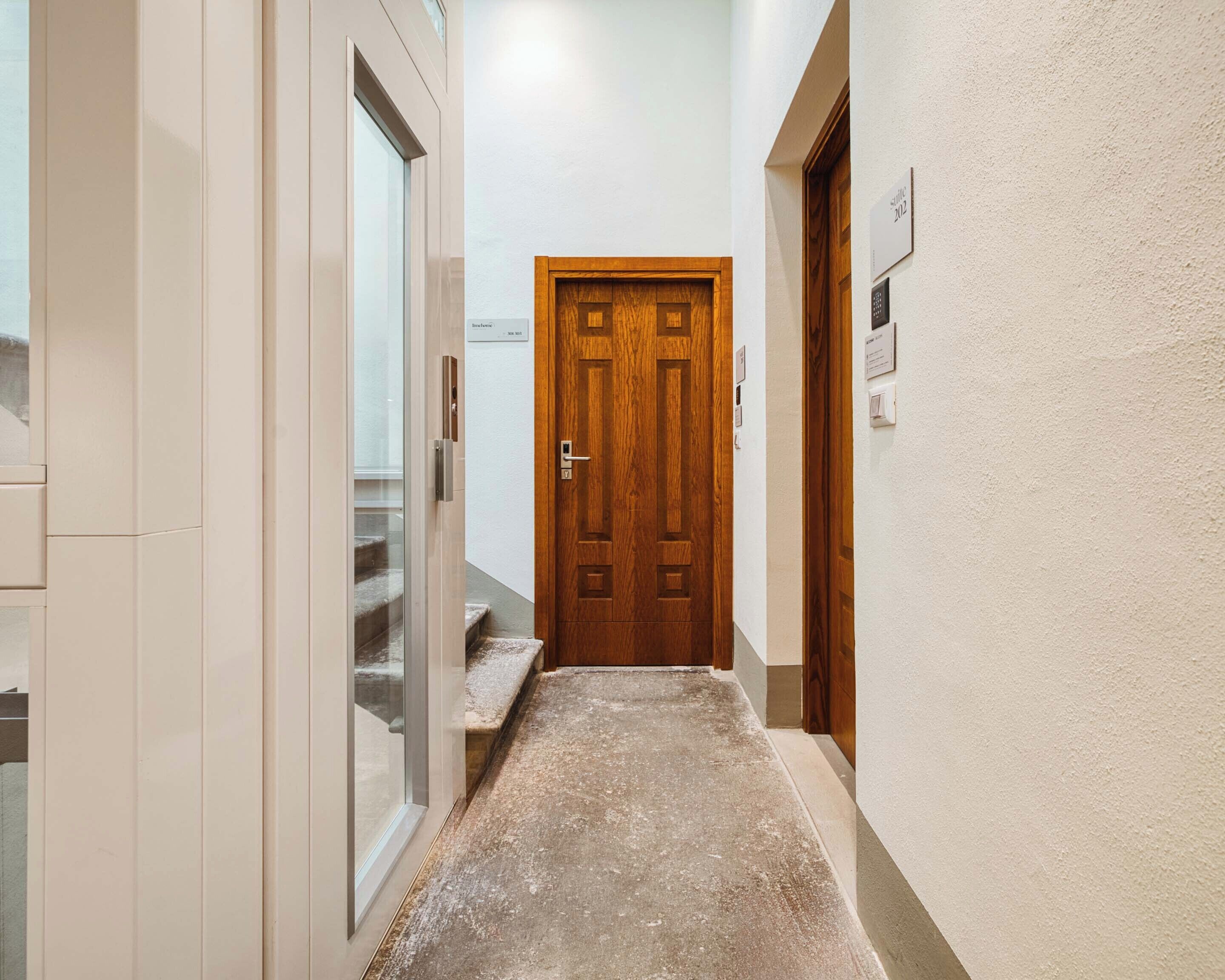 Un couloir étroit, avec une porte en bois au fond, une porte en verre sur la gauche et un mur de couleur claire.