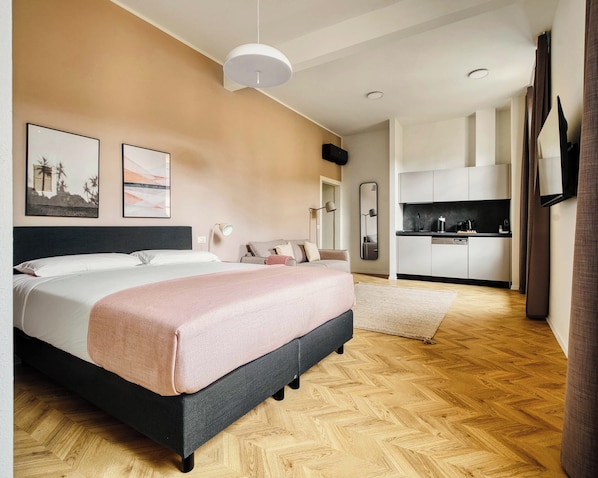 Un dormitorio moderno con cama, mesitas de noche, un espejo y una kitchenette al fondo.