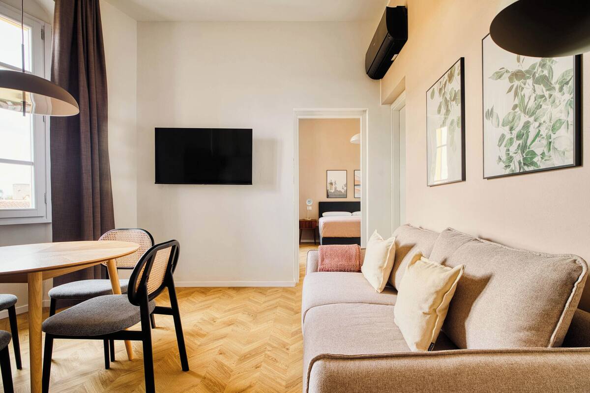 Ein modernes Wohnzimmer mit Holzdielen im Fischgrätmuster, einer Couch, einem Esstisch mit Stühlen, einem Flachbildfernseher und gerahmten Pflanzenbildern an der Wand.