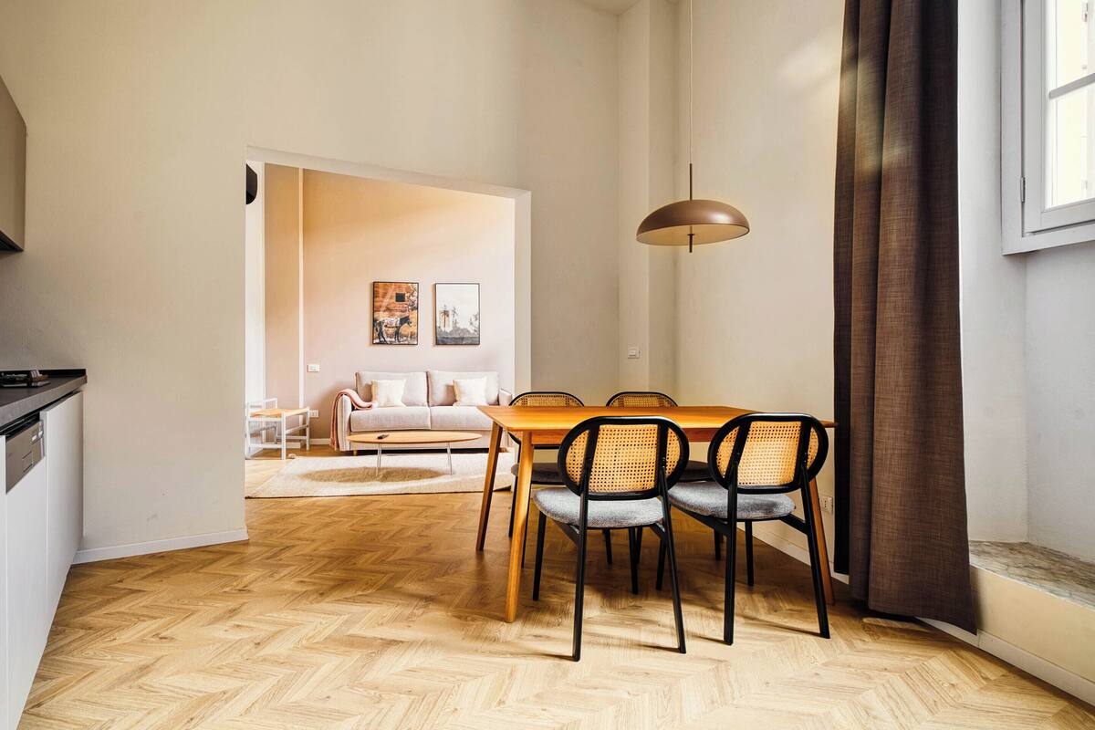 Ein modernes Wohnzimmer mit einer Couch, einem Esstisch mit Stühlen, einer Küchenzeile und einem großen Fenster mit Vorhängen.