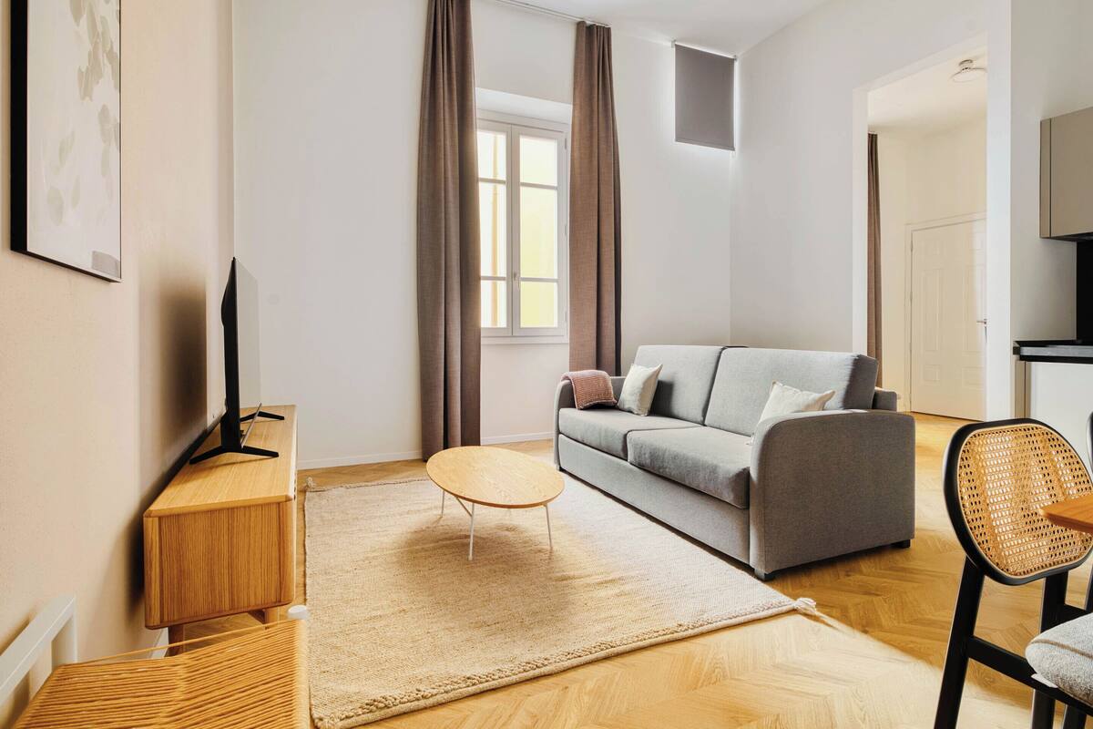 Ein modernes Wohnzimmer mit einer Couch, einem kleinen runden Tisch, einem Fernseher auf einem Holzschrank und einem Korbstuhl.