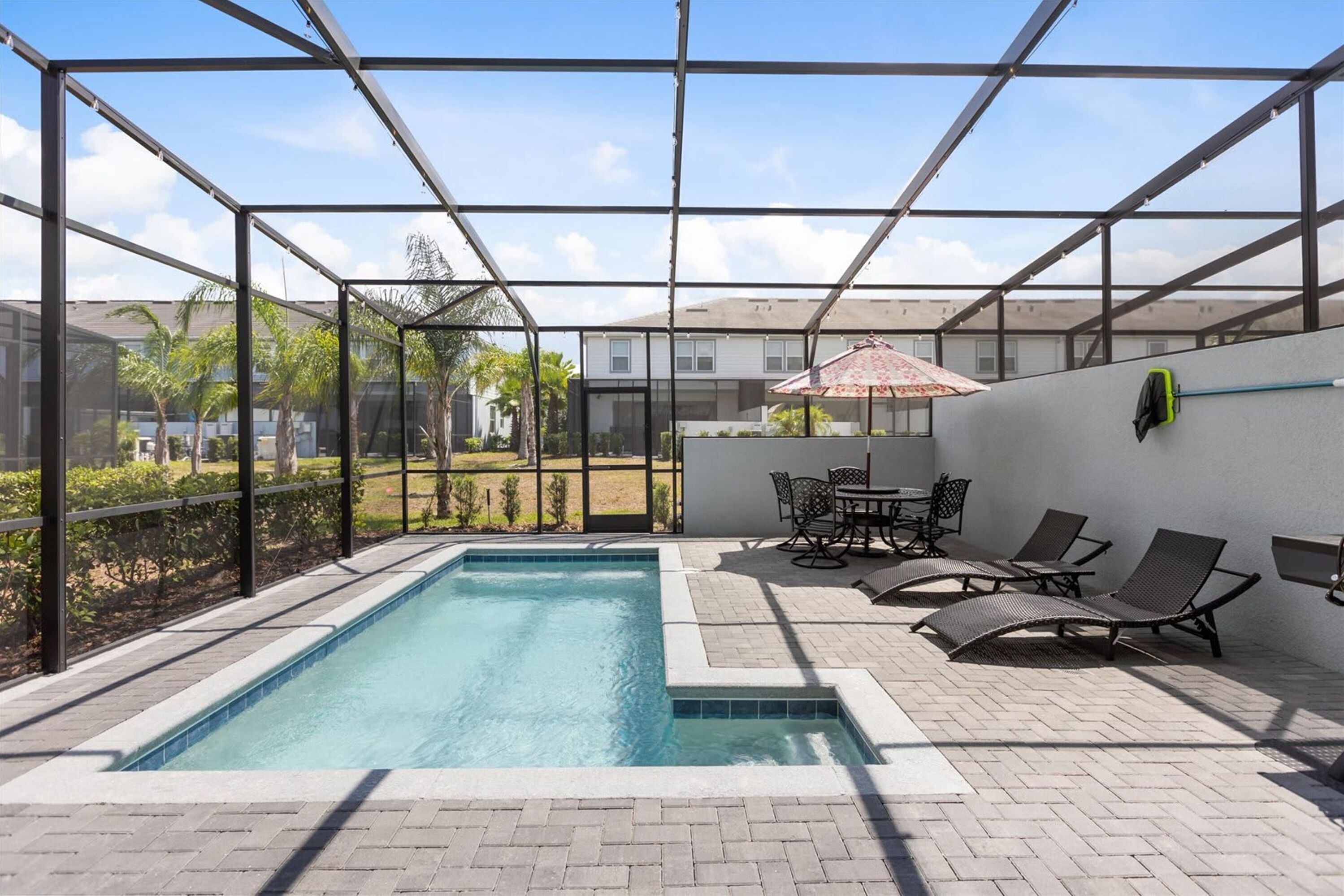 A covered outdoor swimming pool with lounge chairs and a table, surrounded by a metal fence and a view of residential buildings.