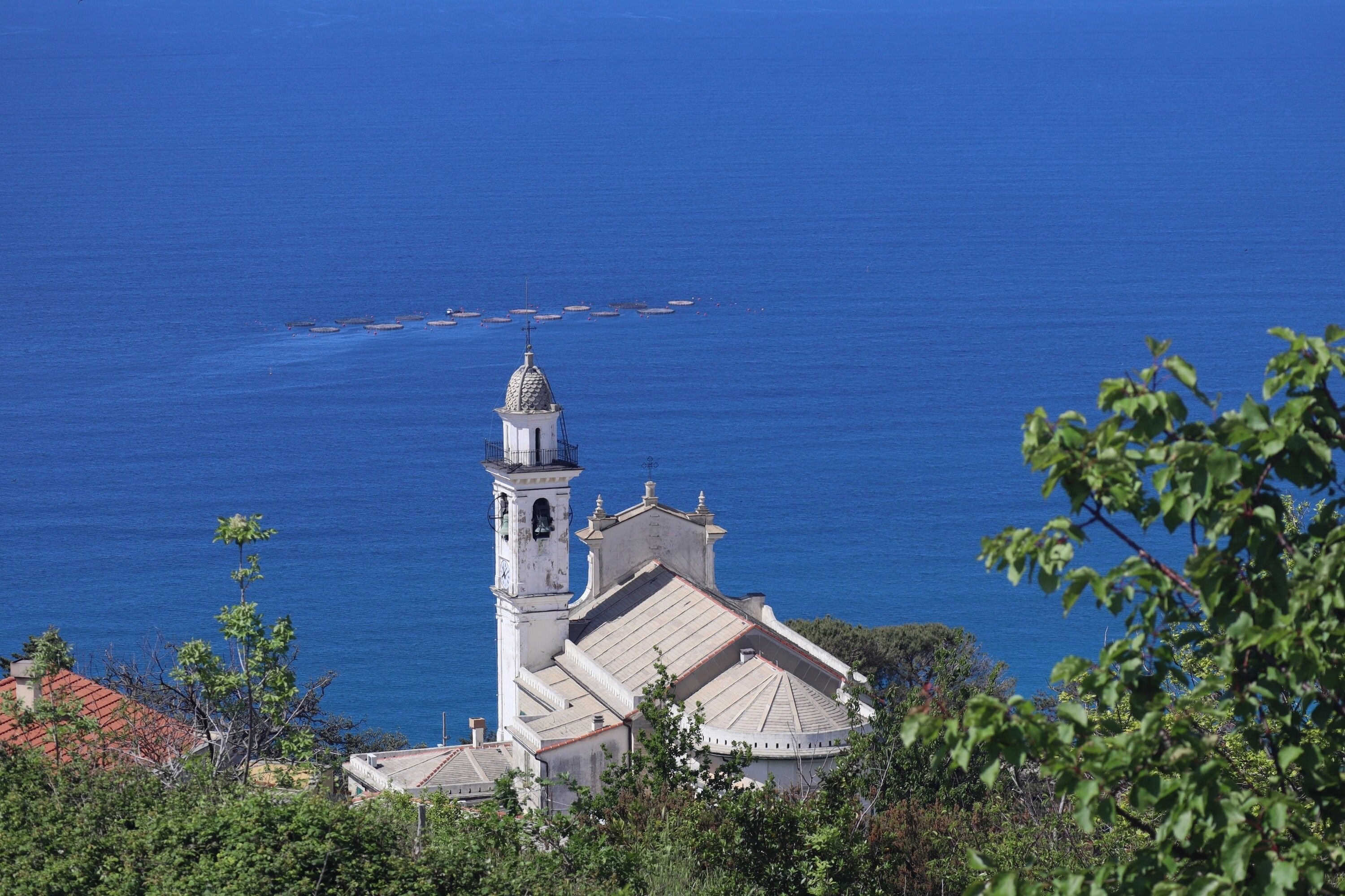 Una iglesia costera con un campanario, rodeada de vegetación y con vistas al mar.