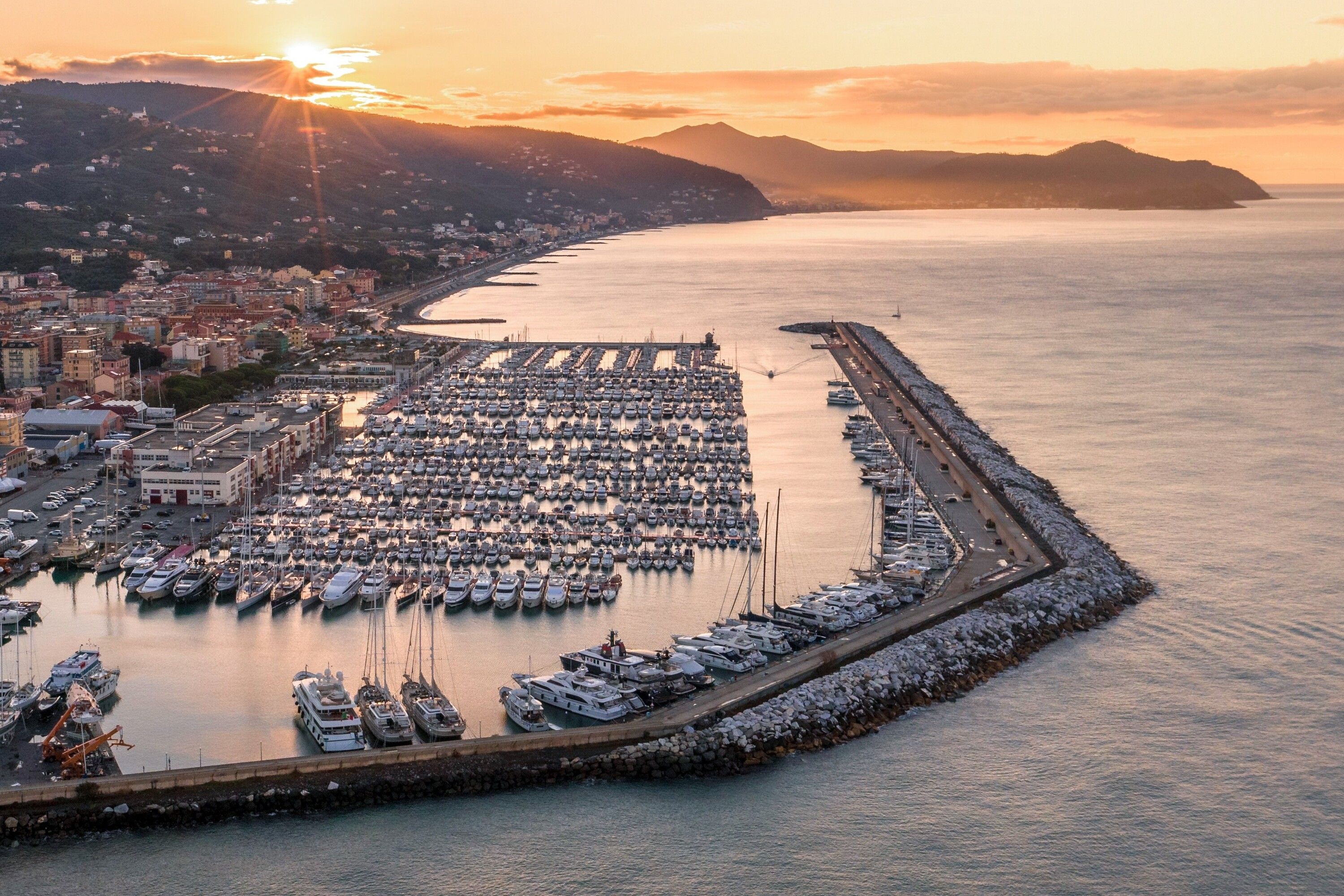 Marina com diversos barcos atracados, um calçadão à beira-mar e um horizonte urbano ao fundo.