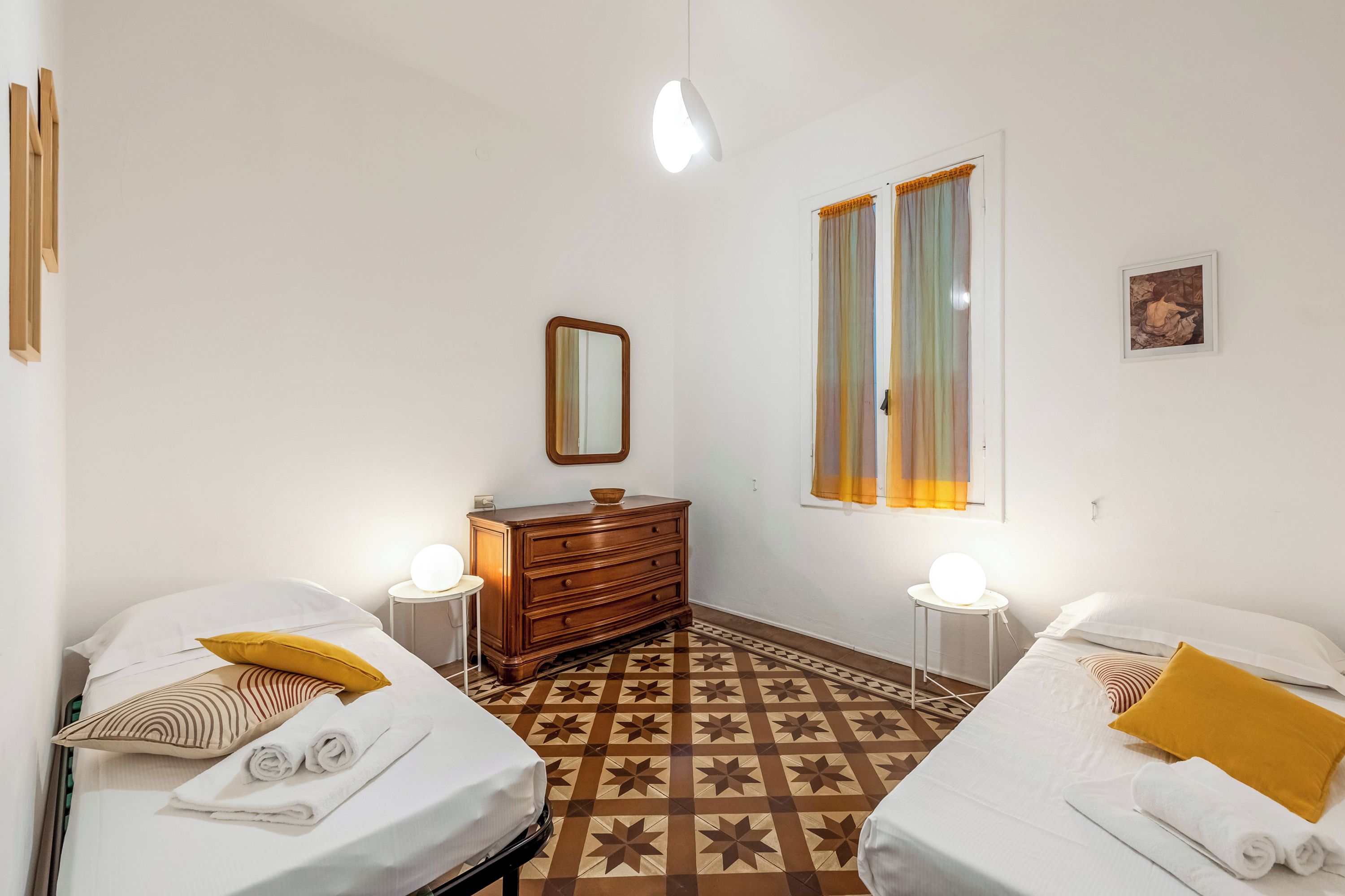 Habitación de hotel con dos camas, un aparador, un espejo, una ventana con cortinas y una mesa redonda con una lámpara, además de un suelo con estampado.