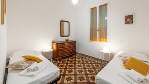 A hotel room with two beds, a dresser, a mirror, a window with curtains, a round table with a lamp, and a patterned floor. - Amiado Apartment With Sea View Balcony (Lavagna)
