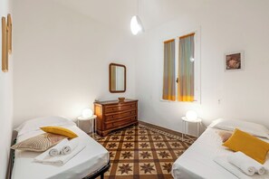 A hotel room with two beds, a dresser, a mirror, a window with curtains, a round table with a lamp, and a patterned floor. - Amiado Apartment With Sea View Balcony (Lavagna)
