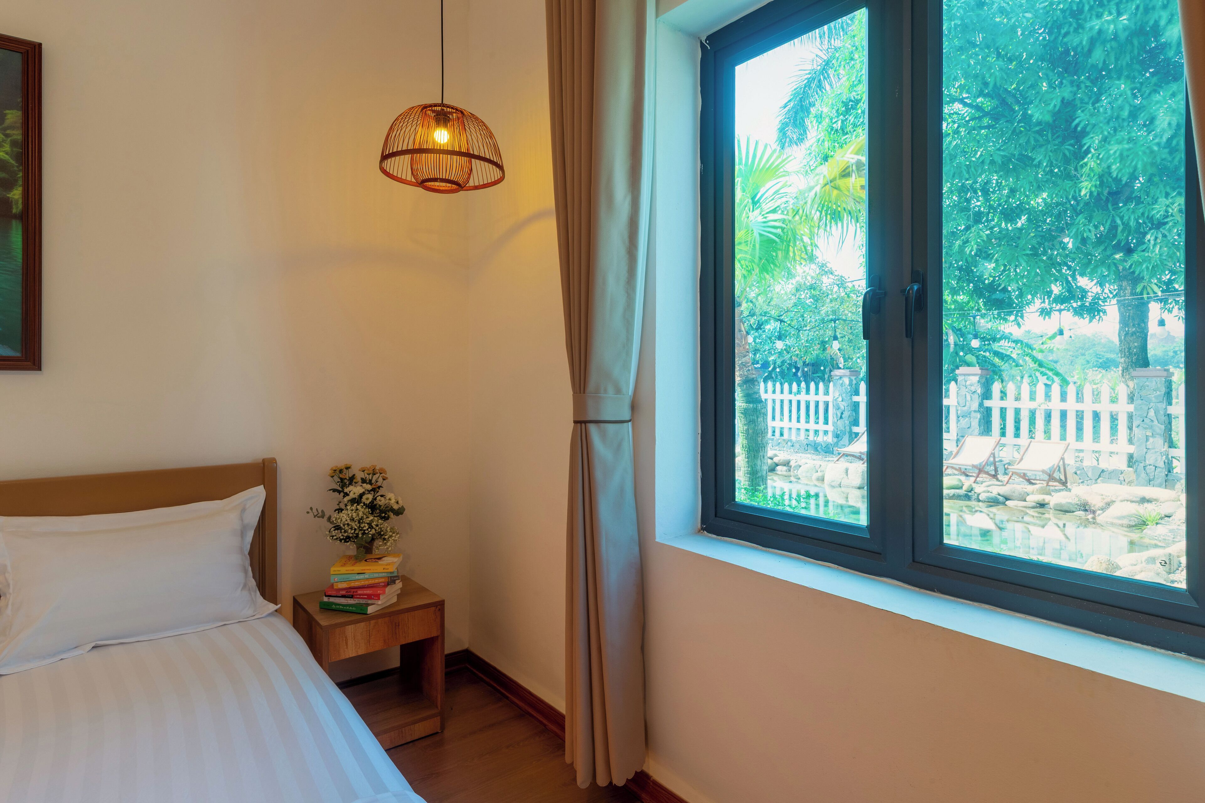 A bedroom with a bed, a bedside table with books, a hanging light fixture, and a window with a view of greenery and a white fence.