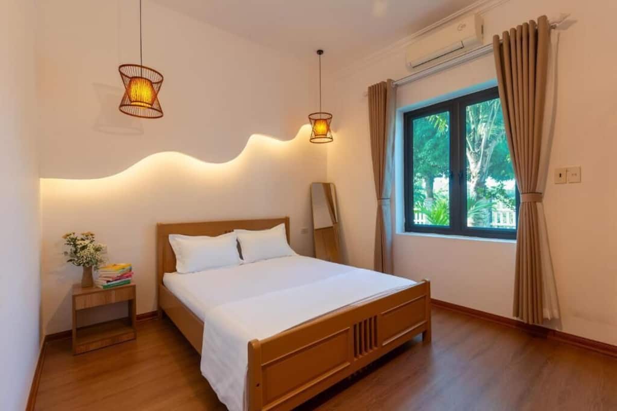 Un dormitorio con cama de madera, mesita de noche con un jarrón de flores y ventana con cortinas.