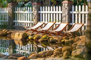 Una hilera de sillones de madera junto a una masa de agua que refleja el paisaje, rodeado de rocas y una valla de piquetes blancos.