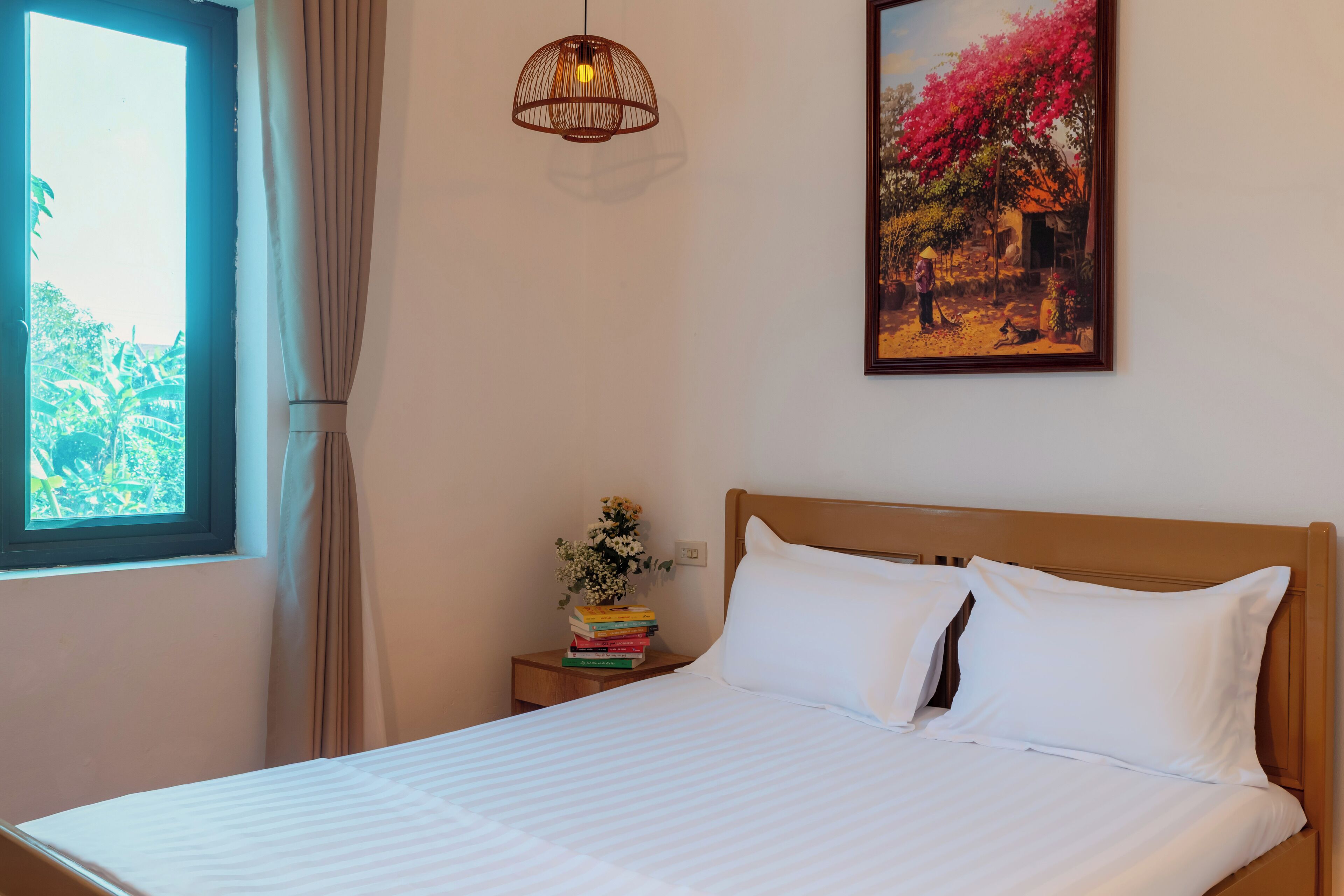 A neatly made bed with white linens, a bedside table with a stack of books and a vase of flowers, a hanging light fixture, and a framed painting on the wall.