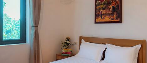 A neatly made bed with white linens, a bedside table with a stack of books and a vase of flowers, a hanging light fixture, and a framed painting on the wall.