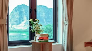 Uma janela com vista para montanhas, um vaso com flores e uma pilha de livros sobre uma pequena mesa de madeira.