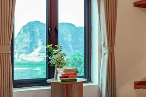 A window with a view of mountains, a vase with flowers, and a stack of books on a small wooden table. - Sol Flow Retreat (Hoa Lu)