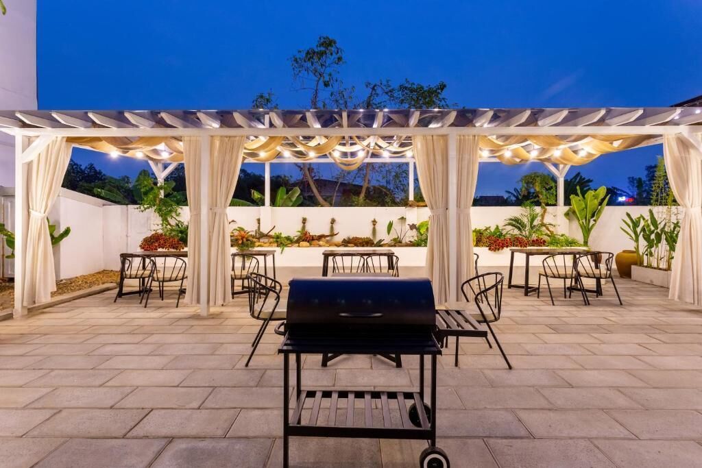 An outdoor patio with a grill, tables, and chairs under a pergola with draped fabric.
