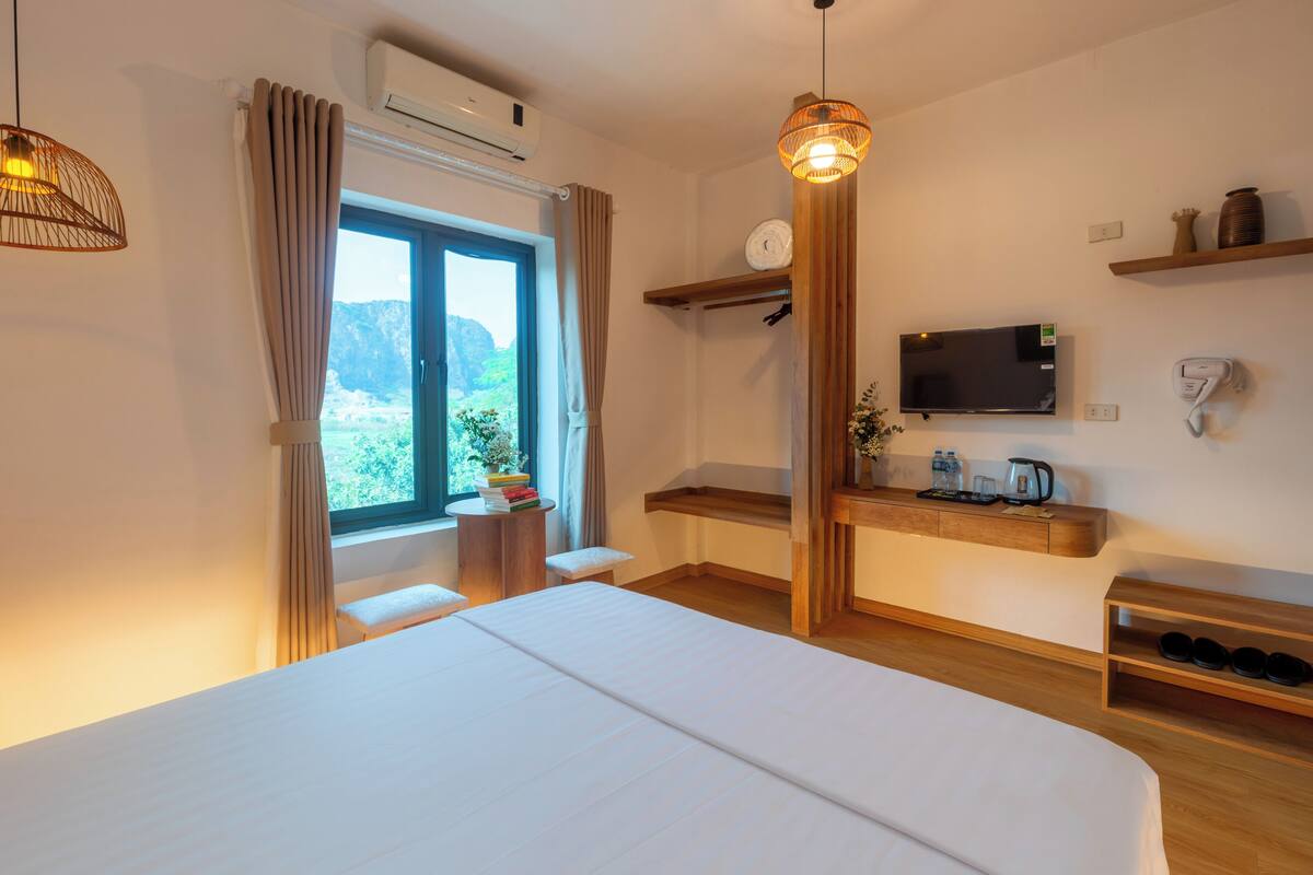 Habitación de hotel con cama, televisión, estante con objetos, ventana con cortinas y aire acondicionado.