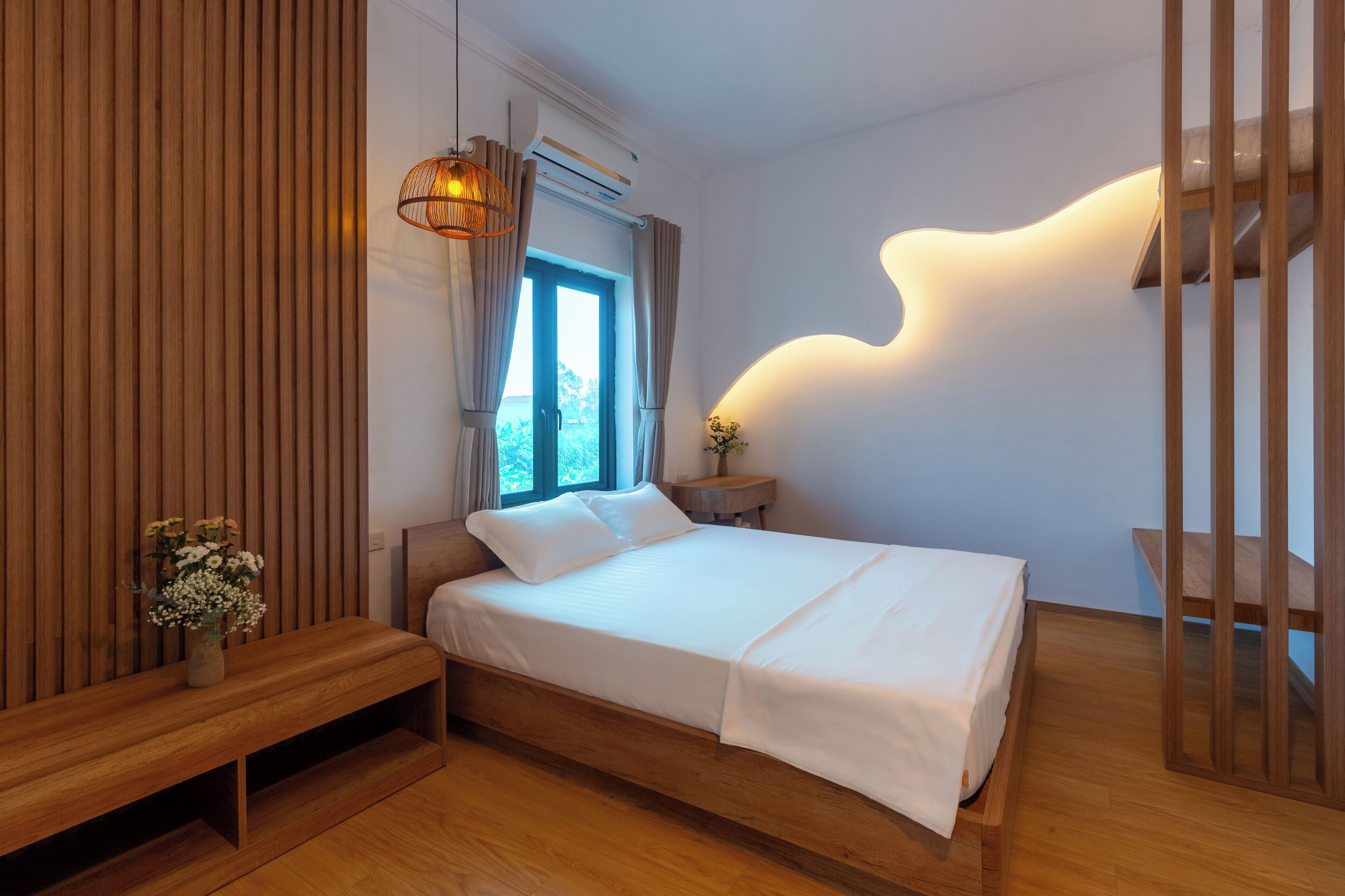 A modern bedroom with a wooden bed, a bench, a bedside table with a vase, and a unique wall light.
