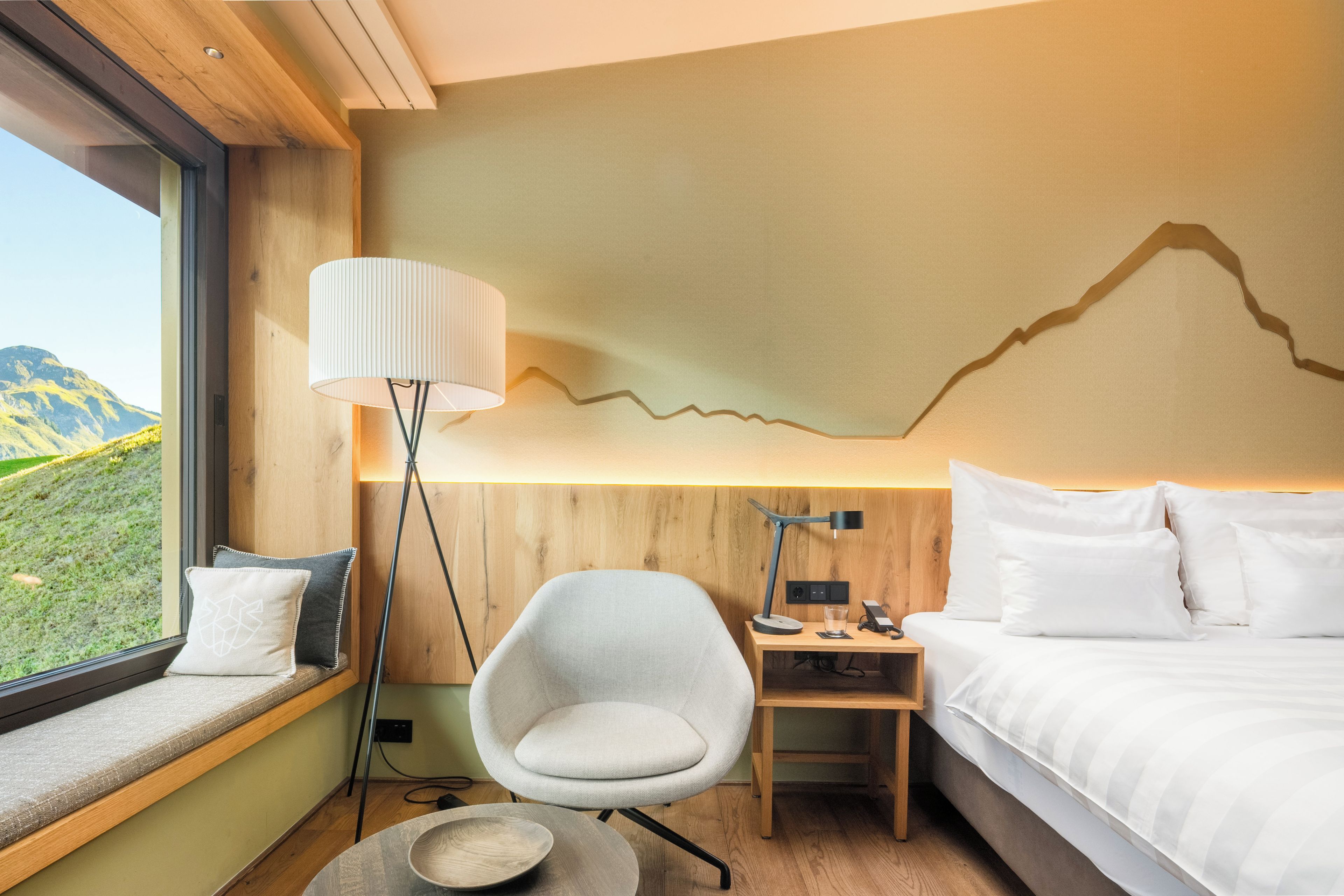 A modern hotel room with a bed, a desk, a chair, and a view of a mountain landscape.