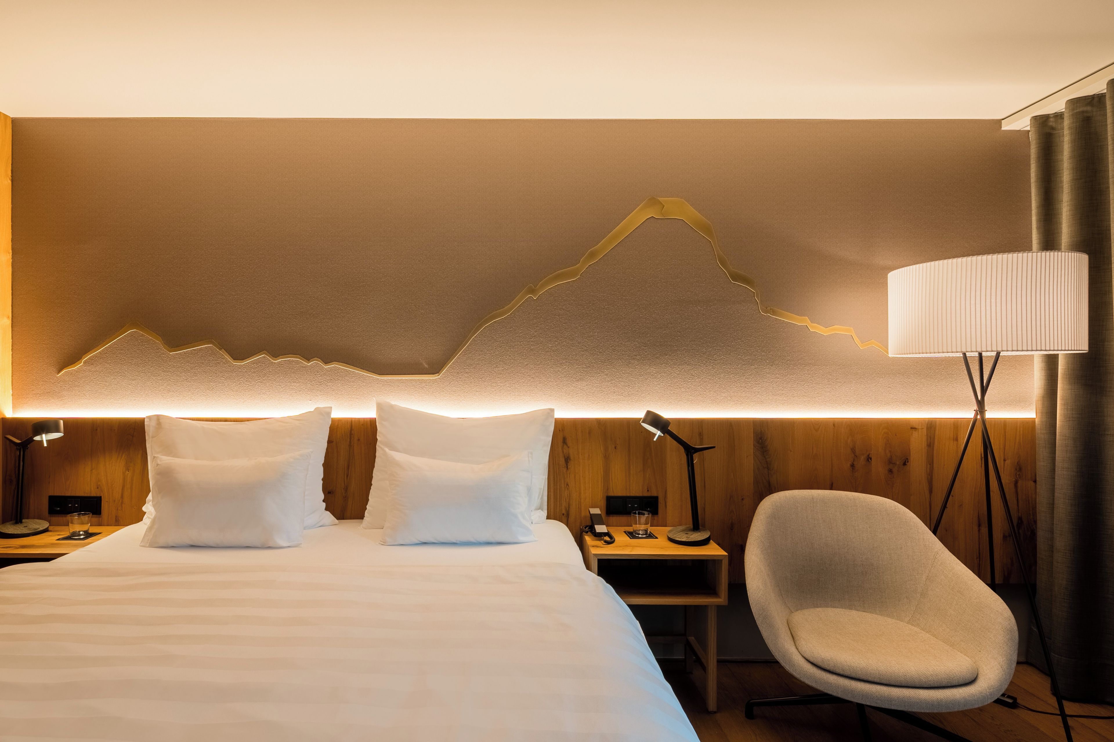 Habitación de hotel con cama, dos almohadas, mesita de noche con lámpara, una silla y una lámpara de pared.