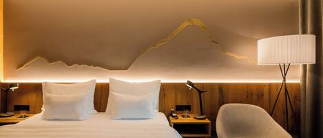 Habitación de hotel con cama, dos almohadas, mesita de noche con lámpara, una silla y una lámpara de pared.