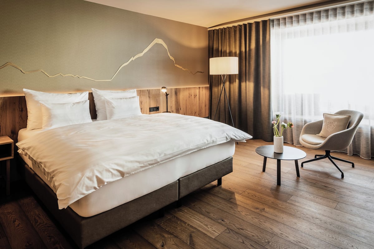 Una habitación de hotel moderna con una cama grande, una mesita de noche, una lámpara, una silla y vistas de la ciudad a través de amplios ventanales.