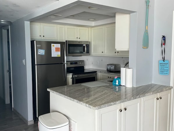A modern kitchen with white cabinets, stainless steel appliances, a granite countertop, and a small island. - Beach   Pool Open  Direct Beachfront Condo  Sunsets (Siesta Key)