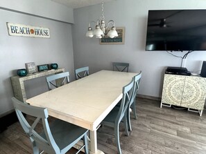 A dining area with a long table, six chairs, a chandelier, a television, and a cabinet with decorative items. - Beach   Pool Open  Direct Beachfront Condo  Sunsets (Siesta Key)