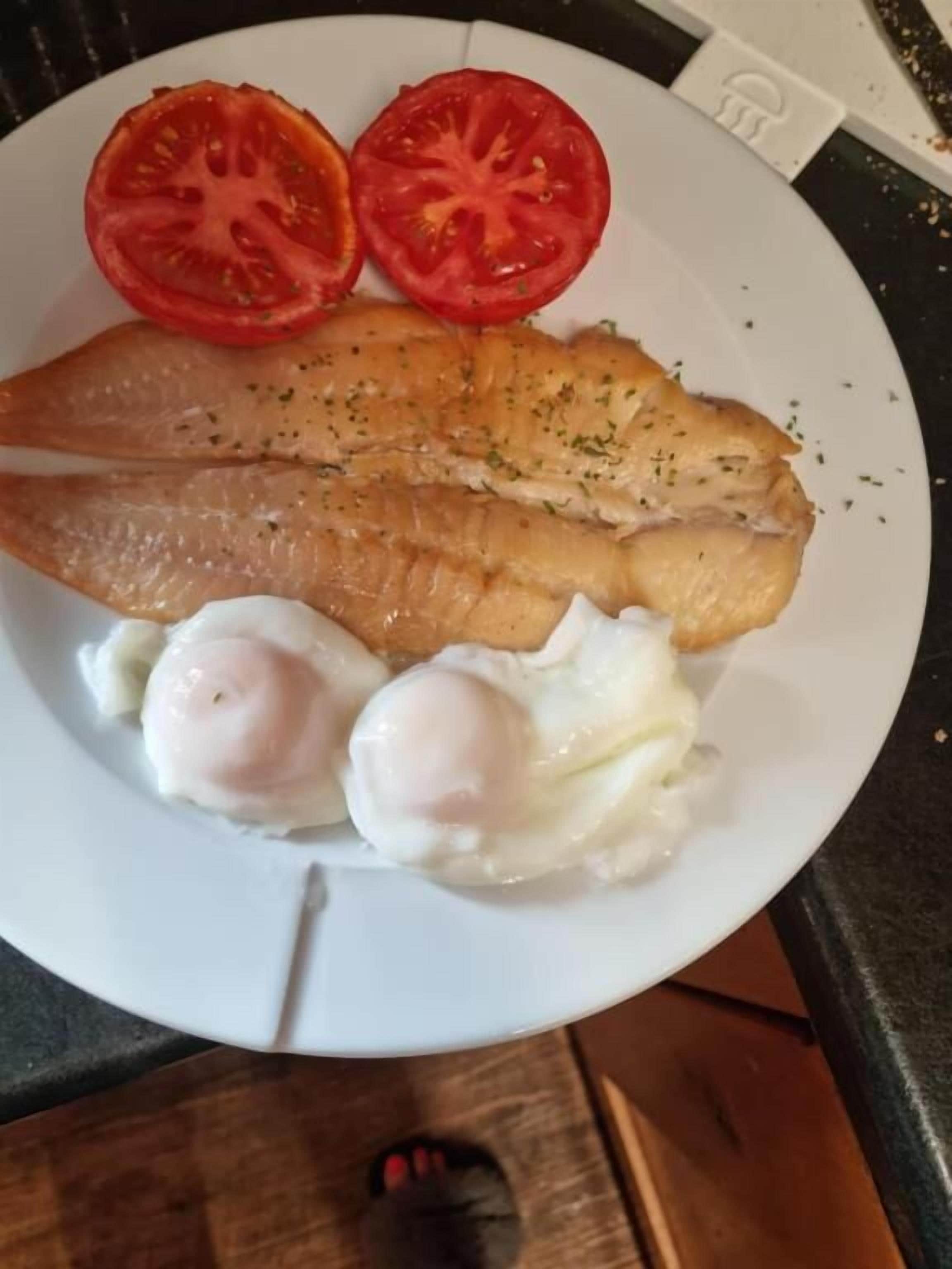 A plate with a piece of fish, two boiled eggs, and two slices of tomato.