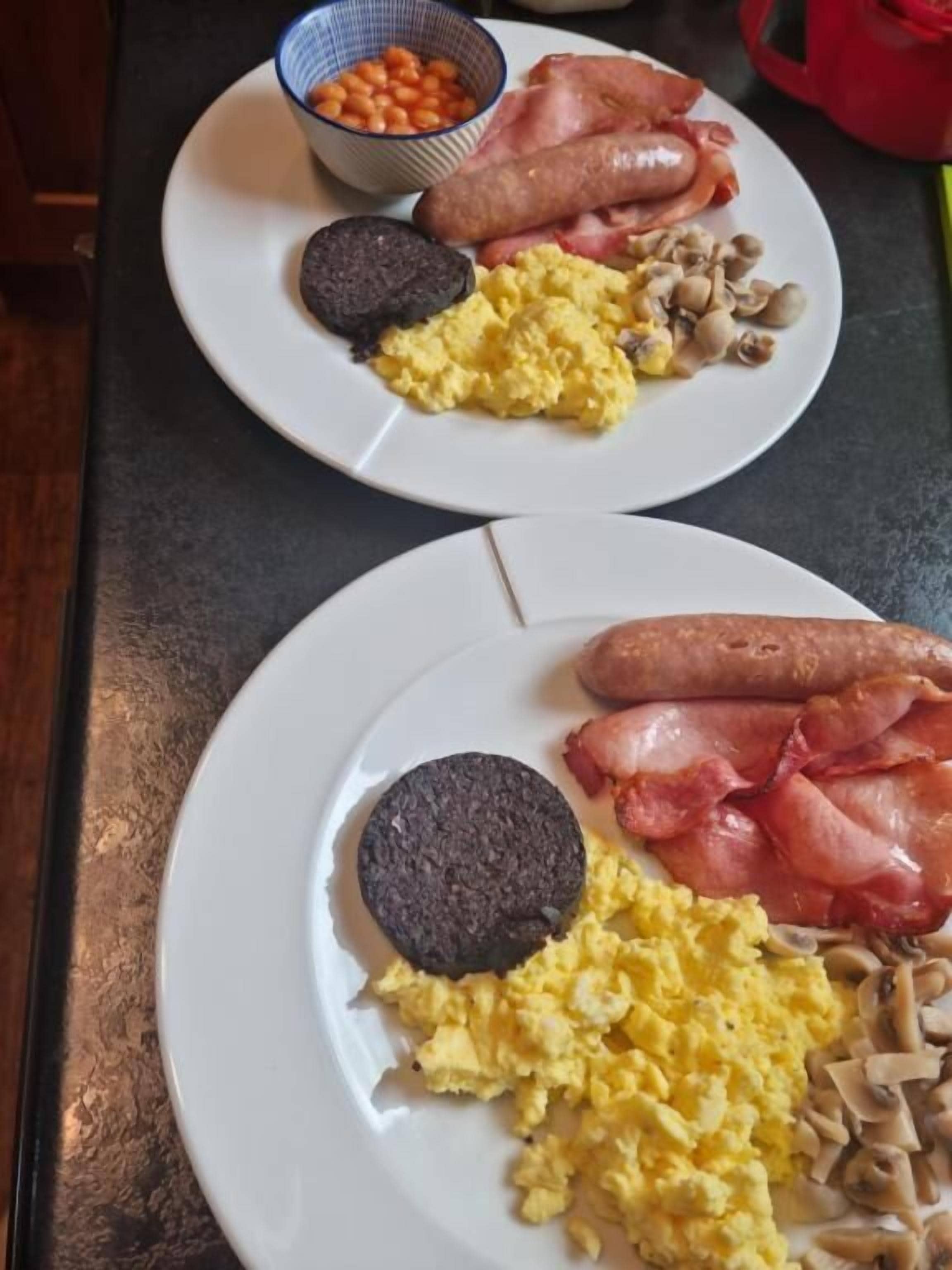 Two plates of breakfast food including sausages, bacon, mushrooms, and scrambled eggs.