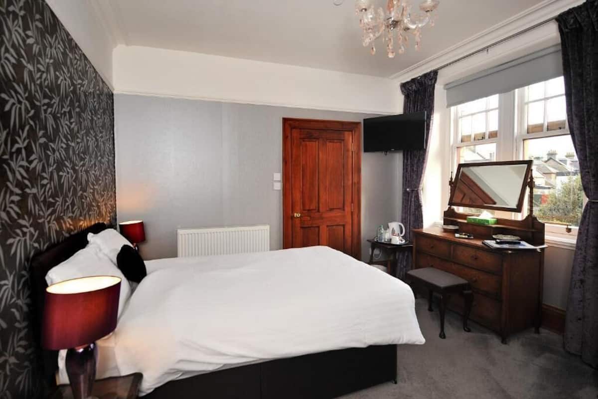 A bedroom with a bed, a desk with a mirror, a radiator, and a window with curtains.