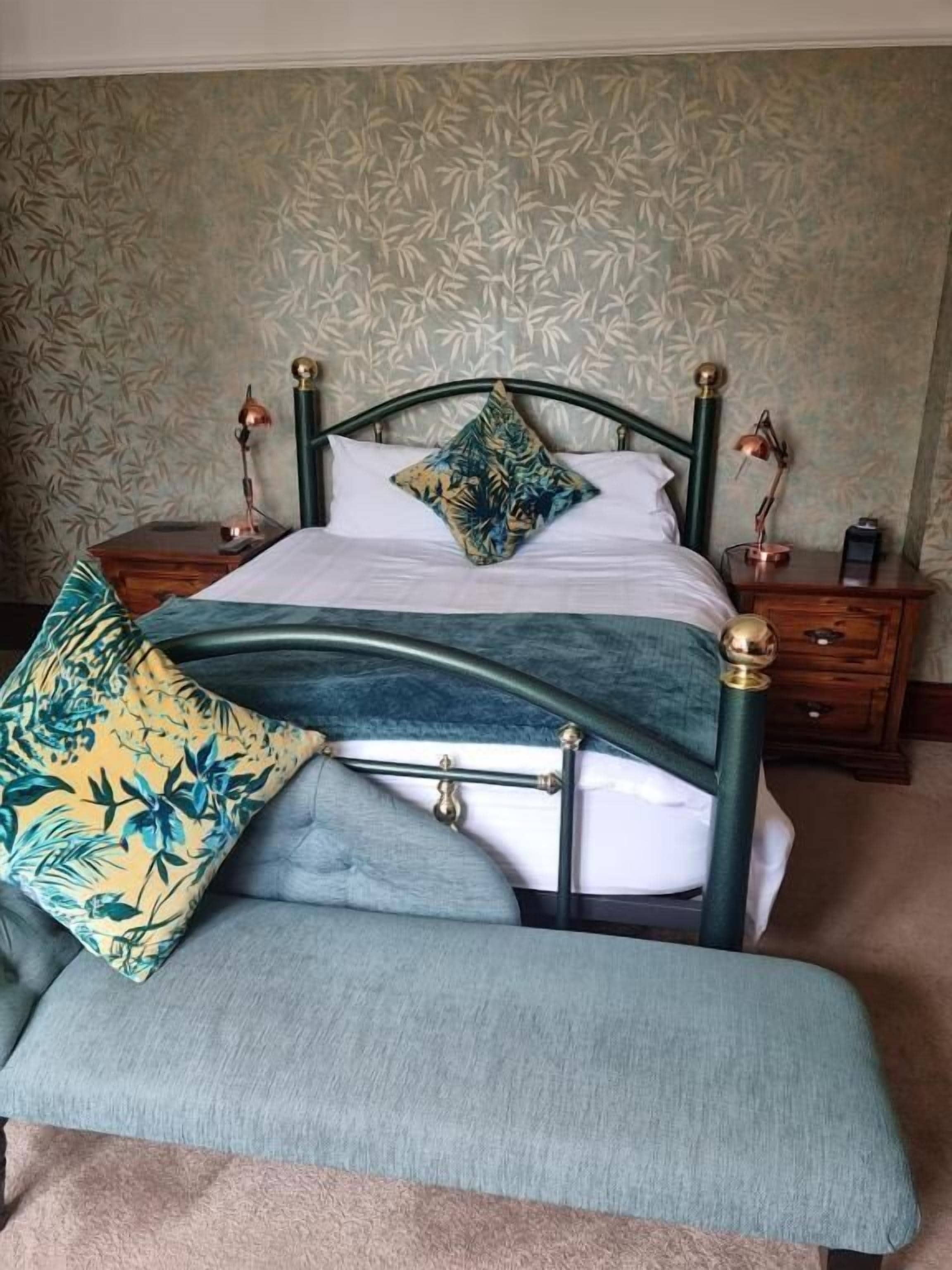 Un lit avec un cadre en métal vert, une literie blanche et un coussin à motifs tropicaux. Il y a un banc avec un motif similaire à celui du coussin.