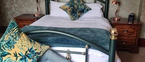 A bed with a green metal frame, white bedding, and a tropical patterned pillow. There is a bench with a similar pattern to the pillow.