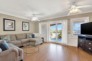 A living room with a beige sofa, a glass coffee table, a ceiling fan, and a TV on a dark wooden dresser. - Coastal Retreat on Tybee Island – Steps to the Beach (Tybee Island)