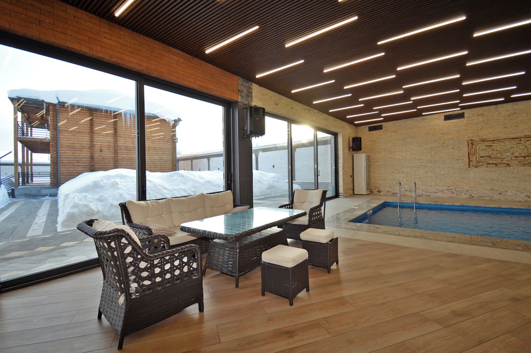 A modern indoor pool area with a glass wall, wicker furniture, and a wooden ceiling.
