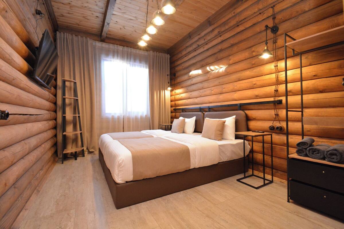 A log cabin bedroom with a large bed, a TV mounted on the wall, a ladder, and a wooden dresser.
