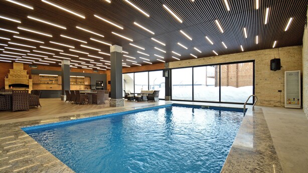 Une piscine intérieure dotée d’un sol carrelé, d’un plafond en bois avec des luminaires linéaires et d’un mur en briques agrémenté d’une cheminée.