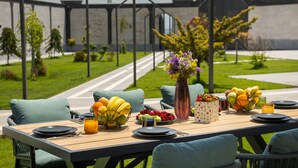 Un espace de restauration en plein air avec une table dressée pour deux personnes, entourée de chaises vertes, d’un vase rempli de fleurs et d’un panier de fruits.