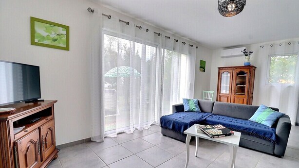 Un salon avec un téléviseur posé sur un meuble en bois, un canapé recouvert d’un plaid bleu et une table basse blanche sur laquelle sont posés des livres.