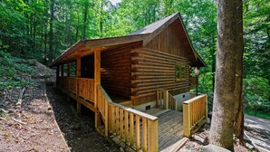 Une cabane en rondins dotée d’un porche et d’une petite terrasse, entourée d’une forêt dense.