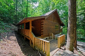 Une cabane en rondins dotée d’un porche et d’une petite terrasse, entourée d’une forêt dense.