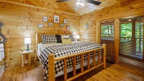 Une chambre à coucher dans une cabane en bois, avec un lit, des lampes de chevet, un ventilateur de plafond et une fenêtre avec des stores.