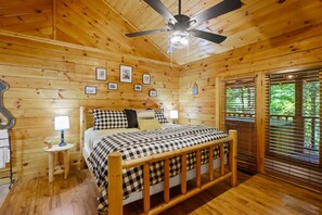 Une chambre à coucher dans une cabane en bois, avec un lit, des lampes de chevet, un ventilateur de plafond et une fenêtre avec des stores.