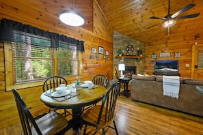 A cozy cabin interior with a dining area, a sofa, a fireplace, and a television. - Location  Log Cabin Hot Tub Fireplace Pool Fishing (Pigeon Forge)