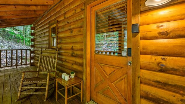 Une cabane en bois avec une grande porte, un fauteuil à bascule et une petite table sur laquelle se trouvent des tasses et une plante.