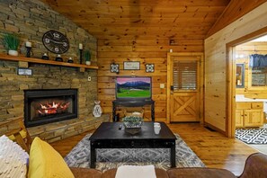 Ein gemütliches Wohnzimmer mit Kamin aus Stein, einem Fernseher und einer Holzbalkendecke.