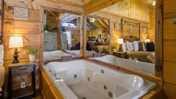 A wooden cabin interior with a large mirror, a bathtub, and a cozy bedroom with a bed and pillows. - Location  Cute Studio Log Cabin Hot Tub Fireplace (Pigeon Forge)